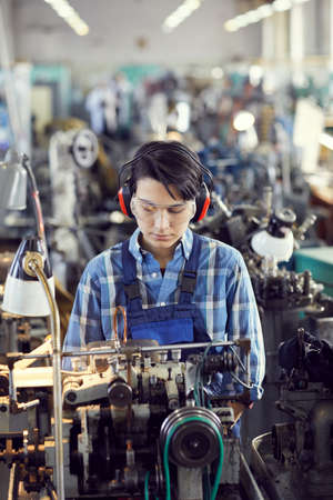 Serious busy young brunette woman in protective goggles standing in industrial shop and preparing manual machine for processing detailの写真素材