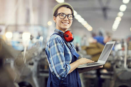 Portrait of cheerful confident hipster female maintenance engineer with ear protectors around neck using laptop at industrial factoryの写真素材