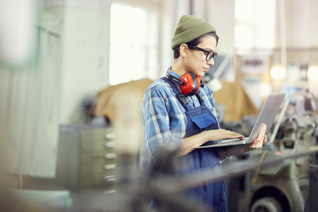 Serious busy young female engineer in hipster hat standing in factory shop and identifying industrial machine errors using laptopの写真素材