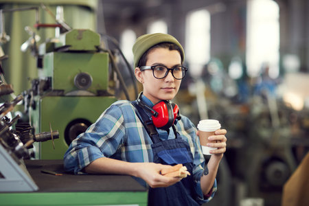 Pensive attractive hipster girl in workwear leaning on factory machine and eating lunch during break