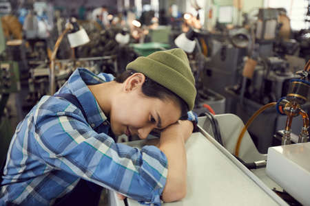 Fatigued young female production worker in green hat tired from work leaning on factory machineの写真素材