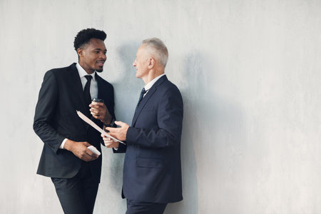 Two multi-ethnic business specialists in formal suits standing at wall and discussing financial...