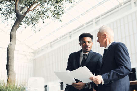 Thoughtful multi-ethnic business specialists standing in light lobby and working with business analysis data while viewing information in papersの写真素材