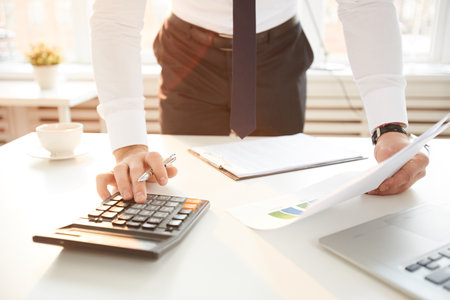 Close-up of unrecognizable businessman in necktie standing at table in office and making calculations while analyzing financial papersの写真素材