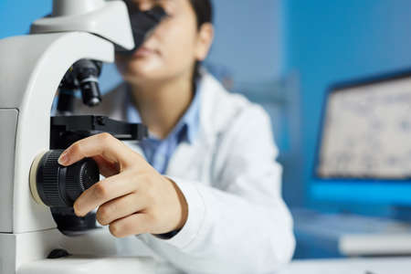 Close-up of female scientist in lab coat sitting at table and using adjustment knob of microscopeの写真素材