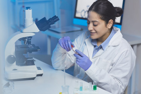 Positive young Indian female medical researcher in lab coat sitting at desk with microscope and...
