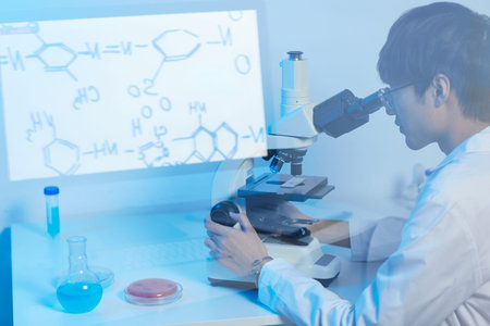 Serious focused modern Asian scientist in glasses sitting at desk in laboratory and using microscope...