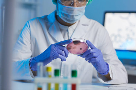 Close-up of busy microbiologist in white coat and rubber gloves sitting at desk and looking at...