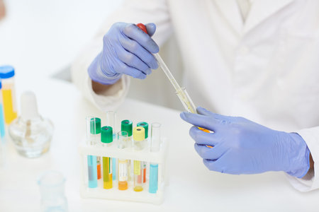 Close-up of unrecognizable scientist in rubber gloves sitting at desk with test tube rack and...