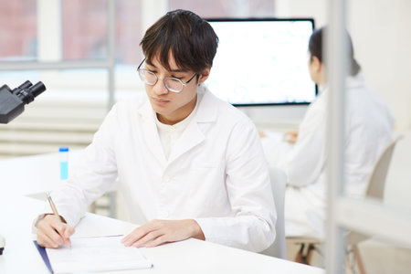 Serious concentrated young Asian lab technician in white coat and eyeglasses sitting at desk and making notes about laboratory researchの写真素材