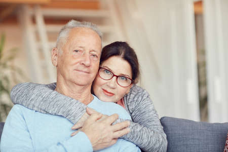 Portrait of happy senior couple resting at home: attractive aged wife embracing husband around neckの写真素材