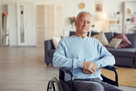 Portrait of serious elderly man with gray hair sitting in wheelchair and holding hands together in modern living roomの写真素材