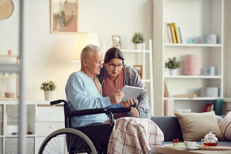 Senior man with legs covered with blanket sitting in wheelchair and showing new mobile app on tablet to surprised wifeの写真素材