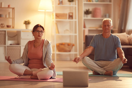 Serious mature couple sitting with crossed legs on mats and holding hands in mudra while meditating...