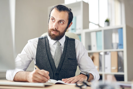 Serious thoughtful young bearded lawyer in gray waistcoat and white shirt sitting at table and...
