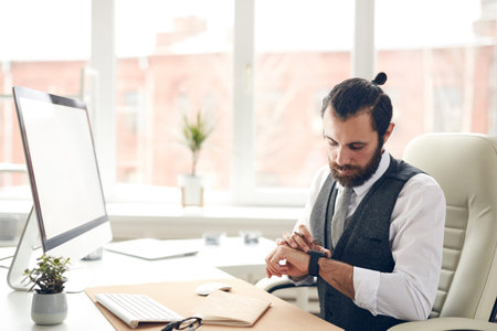 Serious handsome bearded businessman in waistcoat sitting at desk with computer and checking message...