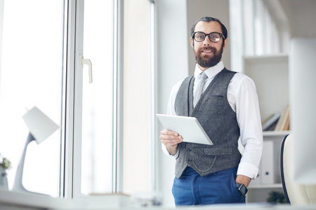 Portrait of smiling modern businessman in eyeglasses standing in office and keeping hand in trousers...