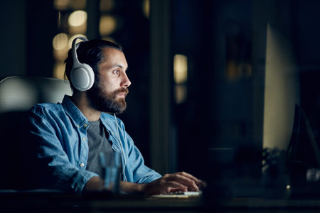 Focused hipster young man with beard sitting at table and listening to audio while working with...