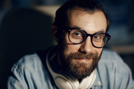 Portrait of content hipster young man with mustache and beard wearing eyeglasses sitting in dark...
