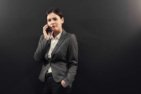 Portrait of content successful young female sales manager in formal jacket standing black background and talking by mobile phoneの写真素材
