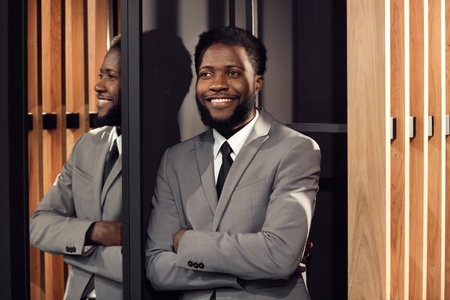 Smiling satisfied young African-American manager with beard leaning on wall and observing business...