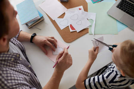Above view of little boy sitting at table and cutting hearts out of paper together with father while they making gift for momの写真素材