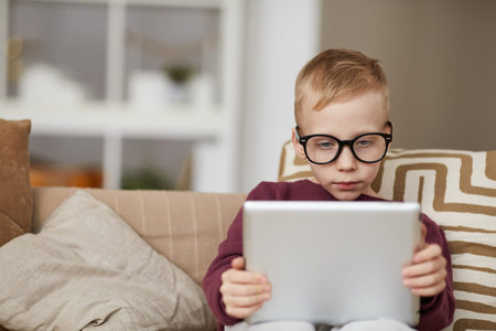 Serious curious little boy in glasses sitting on sofa and using digital tablet while reading online...
