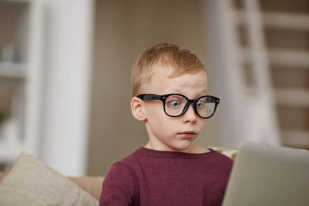 Surprised blond-haired boy in sweater and glasses sitting in living room and seeing internet advertising messageの写真素材