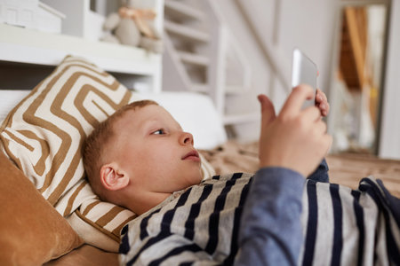 Close-up of serious addicted kid lying on bed and using digital tablet while watching movie on...