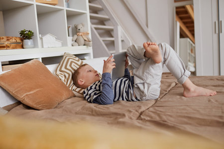 Serious relaxed boy in casual outfit lying on bed and using modern tablet while watching video on...