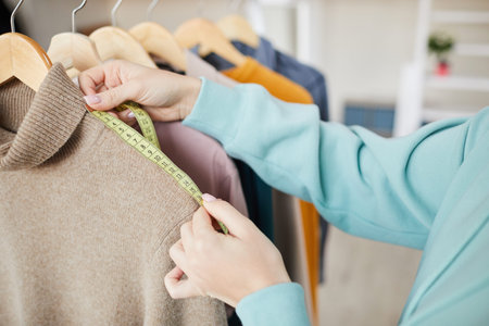Close-up of unrecognizable female dressmaker in sweatshirt standing at clothes rack and using...