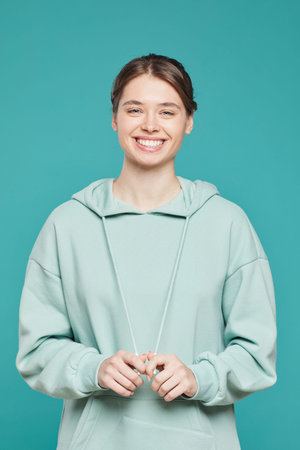 Portrait of positive hipster girl in hoodie standing against teal background and pulling strings of...