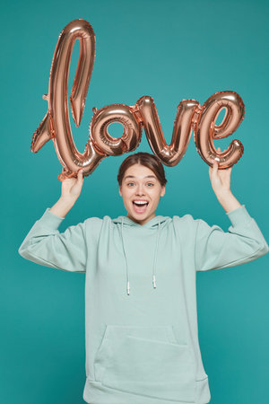 Portrait of positive playful girl in stylish hoodie standing against bright background and holding...
