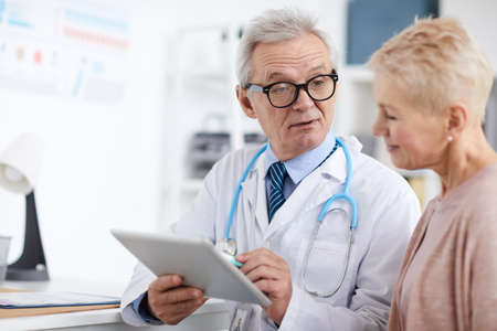 Serious wrinkled senior doctor in lab coat sitting at table and using digital tablet with online patients records while giving recommendations to mature womanの写真素材