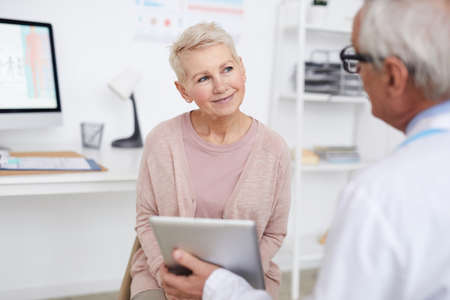 Over shoulder view of elderly doctor using tablet while showing tests results to senior patient at appointmentの写真素材