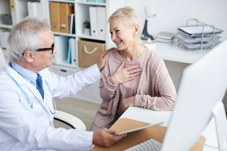 Happy senior female patient with short hair sitting at table in doctors office and holding hand on chest while thanking doctor for good treatment and supportの写真素材
