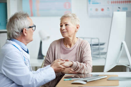 Senior doctor in eyeglasses sitting at table and holding hand of mature female patient while giving hope and comfort to herの写真素材
