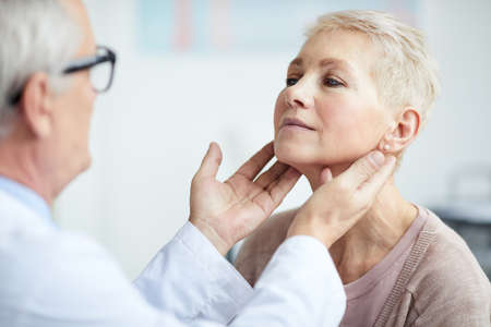 Over shoulder view of senior endocrinologist touching neck of mature female patient while examining her thyroidの写真素材