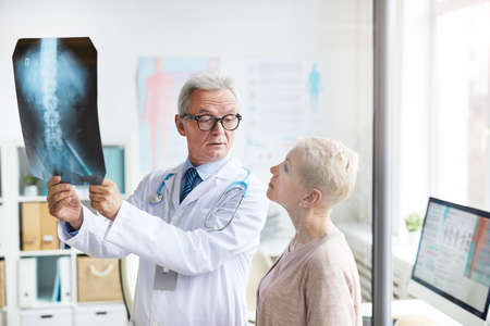 Frowning handsome aged doctor in eyeglasses holding x-ray image against light while explaining spinal problems to senior female patientの写真素材