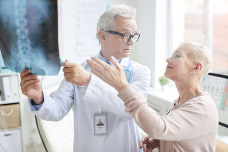 Attractive blond-haired mature lady pointing at core x-ray image while discussing her pain with doctorの写真素材