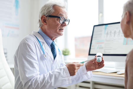 Serious wrinkled physician in glasses pointing with pen at pill bottle while advising patient to take medicineの写真素材