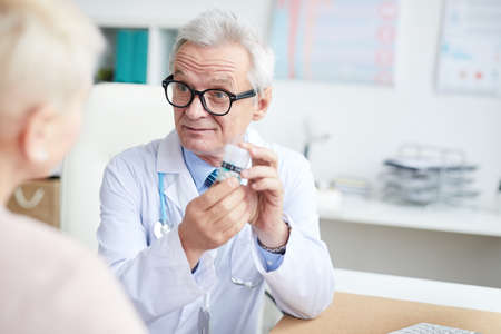 Content senior physician in eyeglasses frowning forehead while prescribing pills to patient at consultationの写真素材