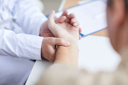 Close-up of unrecognizable male physician in lab coat sitting at desk and keeping finger on patients wrist while analyzing his pulse beatsの写真素材