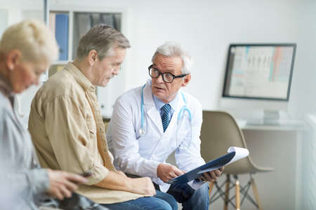 Serious senior male doctor in white coat and eyeglasses holding clipboard with medical card and answering patients questionsの写真素材