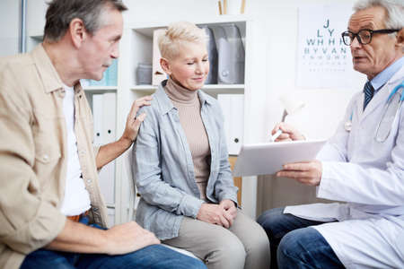 Content mature wife in casual outfit supported by husband attentively listening to doctors advices while her attending doctor reporting good news at appointmentの写真素材