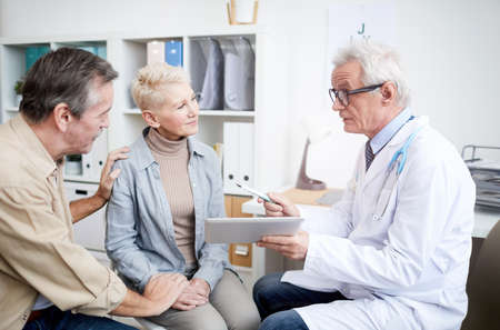 Serious high-qualified cancer doctor in glasses showing medical records on tablet and giving advice to mature woman with serious disease, her husband holding her hand as symbol of supportの写真素材