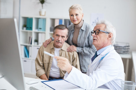 Serious senior male doctor in eyeglasses sitting at desk and using computer while showing test results to man supported by wife at consultationの写真素材