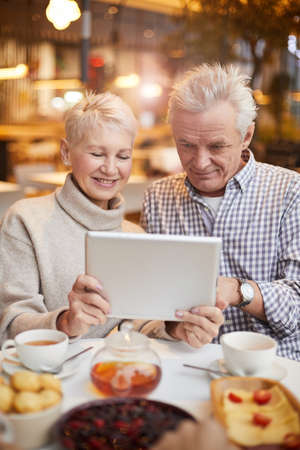 Smiling senior couple in casual outfits sitting at table with tea and desserts and using tablet while watching movie onlineの写真素材