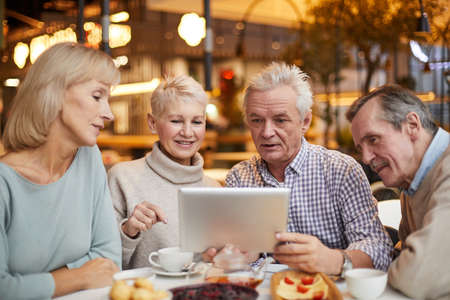 Group of content curious senior friends in casual outfits sitting at table in cafe with wi-fi and surfing net on tablet togetherの写真素材