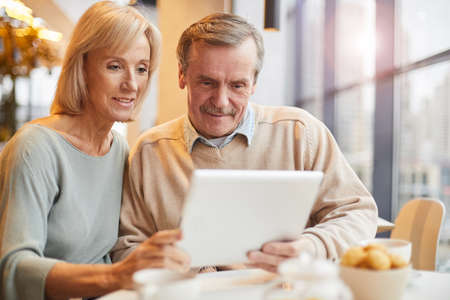 Smiling senior couple in casual clothing sitting at table and using modern device while surfing net together in cafeの写真素材